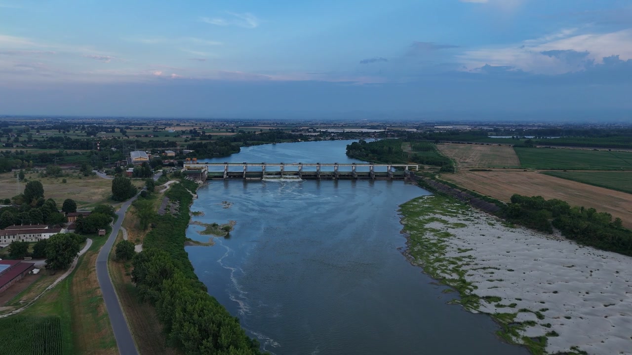 La diga di Isola Serafini attraversa il fiume Po vicino a Monticelli d'Ongina, Piacenza, con sedimenti fluviali esposti, campi agricoli e strade rurali che circondano la diga idroelettrica, ripresa aerea con drone