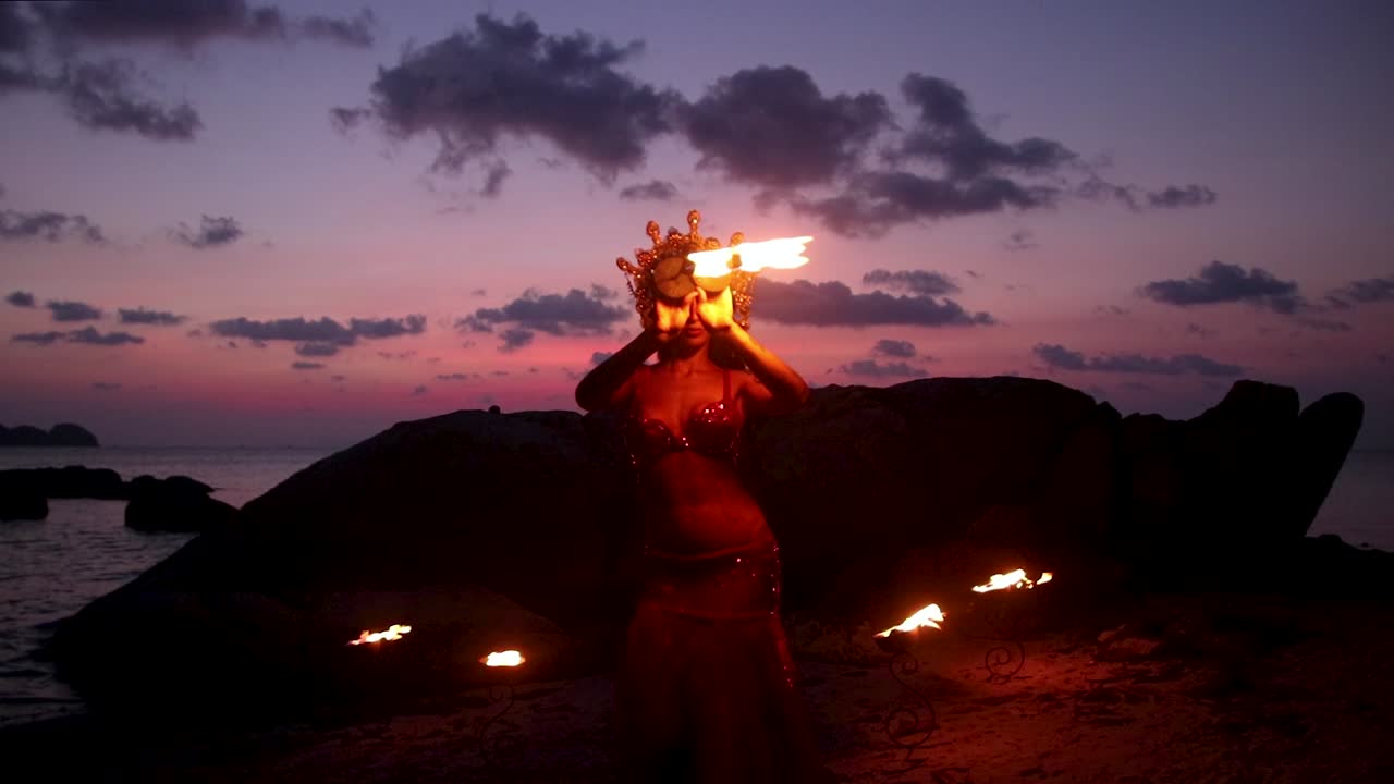 Slow motion of erotic and sensual belly dancer in red dress performing with fire palm torch props, on the beach during golden hour