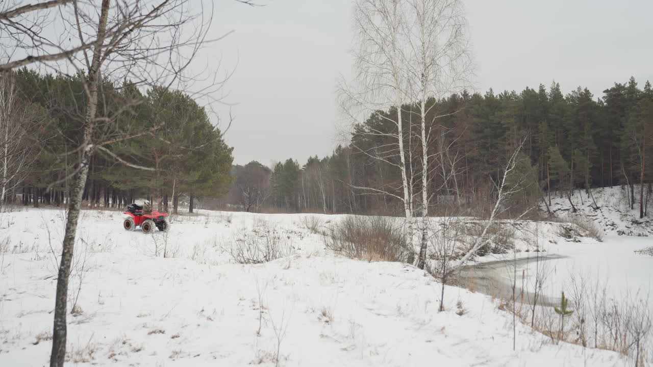 ATV rosso che attraversa un panorama di sponda fluviale innevata. Ampia inquadratura paesaggistica con un pilota in lontananza che naviga lungo la palude ghiacciata e il bordo dello stagno. Lento movimento panoramico, calma luce invernale, con uno sfondo di cresta di pini. Esplorazione.