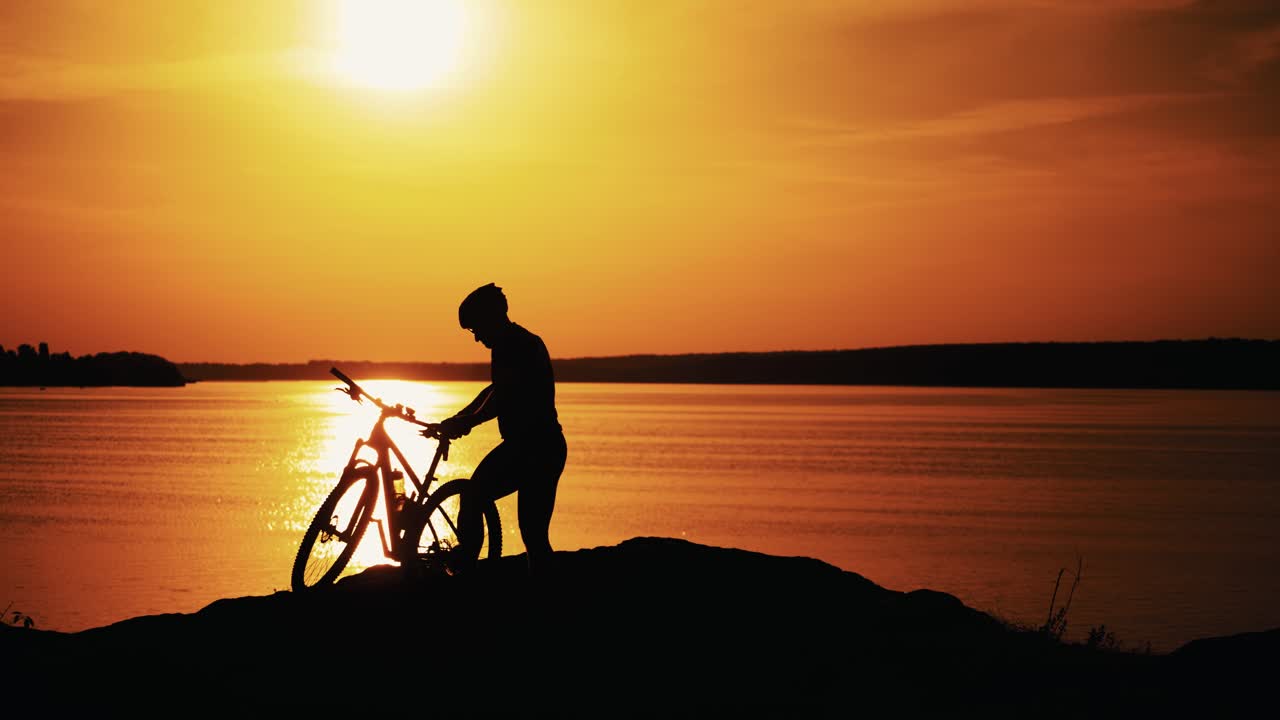 Professional male biker gets off the bike near the river at a beautiful sunset. Silhouette of a sporty cyclist in helmet on the evening water background at orange setting sun.