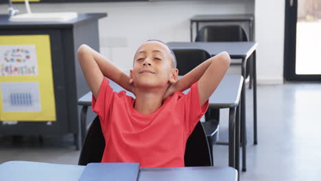 biracial boy with a shaved head, wearing a red shirt, relaxes in a classroom at school