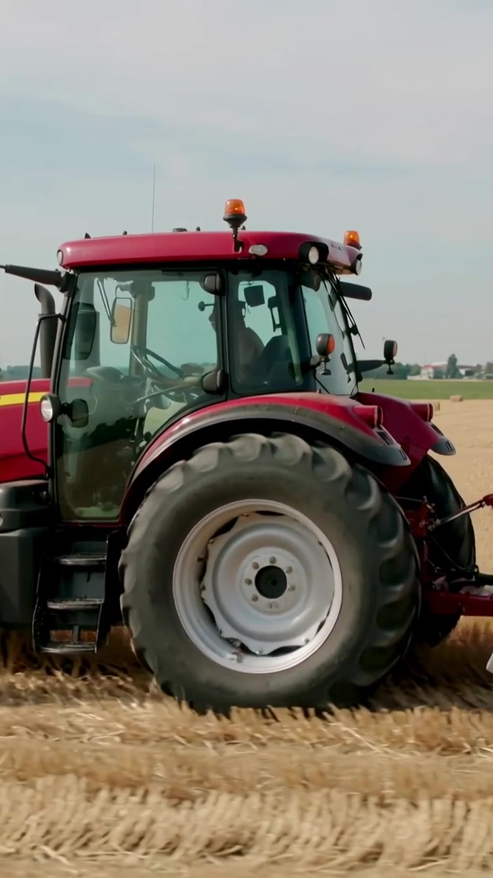 Video verticale: Trattore rosso in movimento, conducente con cappellino che aziona una pressa per balle attraverso un campo tagliato, formando balle