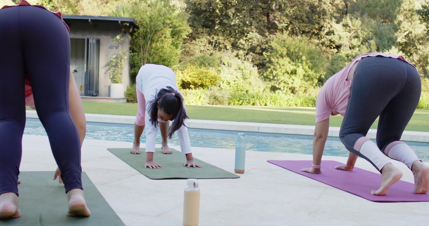 Diverse yogi donne che passano dalla posizione del piegamento in avanti in piedi, posizionando le mani sui tappetini per approfondire la posa a bordo piscina