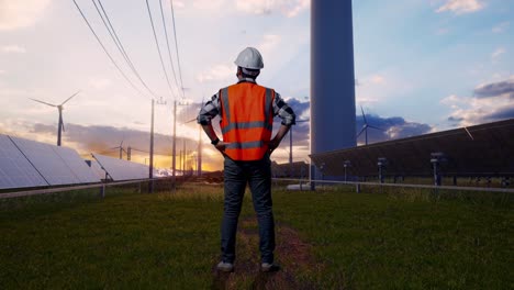 full body back view of a male engineer wearing safety helmet looking around while standing with arms akimbo with solar panel and wind turbines