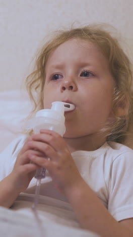 upset little blonde girl undergoes medical procedure with inhaler lying in bed with blanket in light room