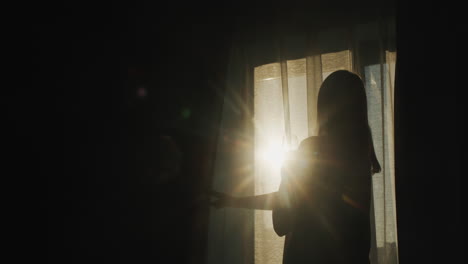 silhouette of a woman who opens blackout curtains. it is illuminated by the bright sun from the window. back view