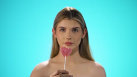 static medium shot of a young pretty blonde woman holding a lollipop to the camera and then taking it with relish while she licks it and has a smile on her lips in front of turquoise background