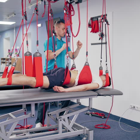 physiotherapist is rising his client man with a special suspension rope system. young man is hanging by rope system in a rehabilitation center. instructor helps his patient to recover.