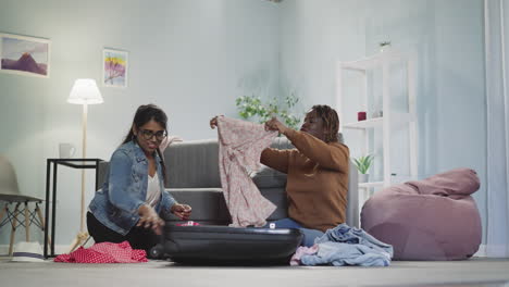 happy multiracial friends put dresses into tourist suitcase