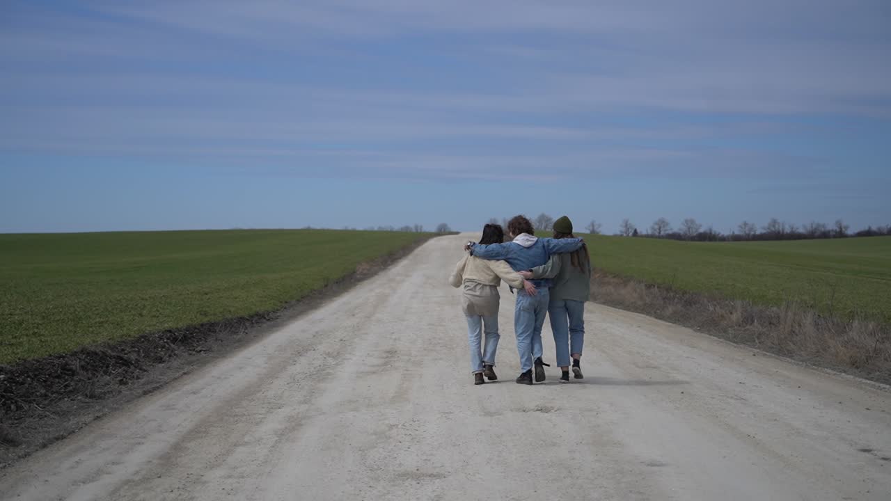 Two young girls and a young boy walk backwards along a road in each other's arms.