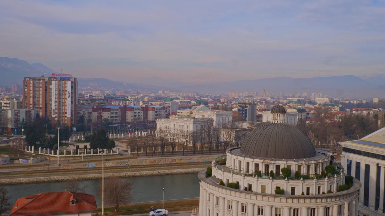 Vista aérea de alto ângulo do horizonte da cidade de Skopje na Macedônia do Norte, mostrando o aterro do rio Vardar, um edifício neoclássico com cúpula, arranha-céus residenciais e montanhas distantes sob um céu de inverno nebuloso.