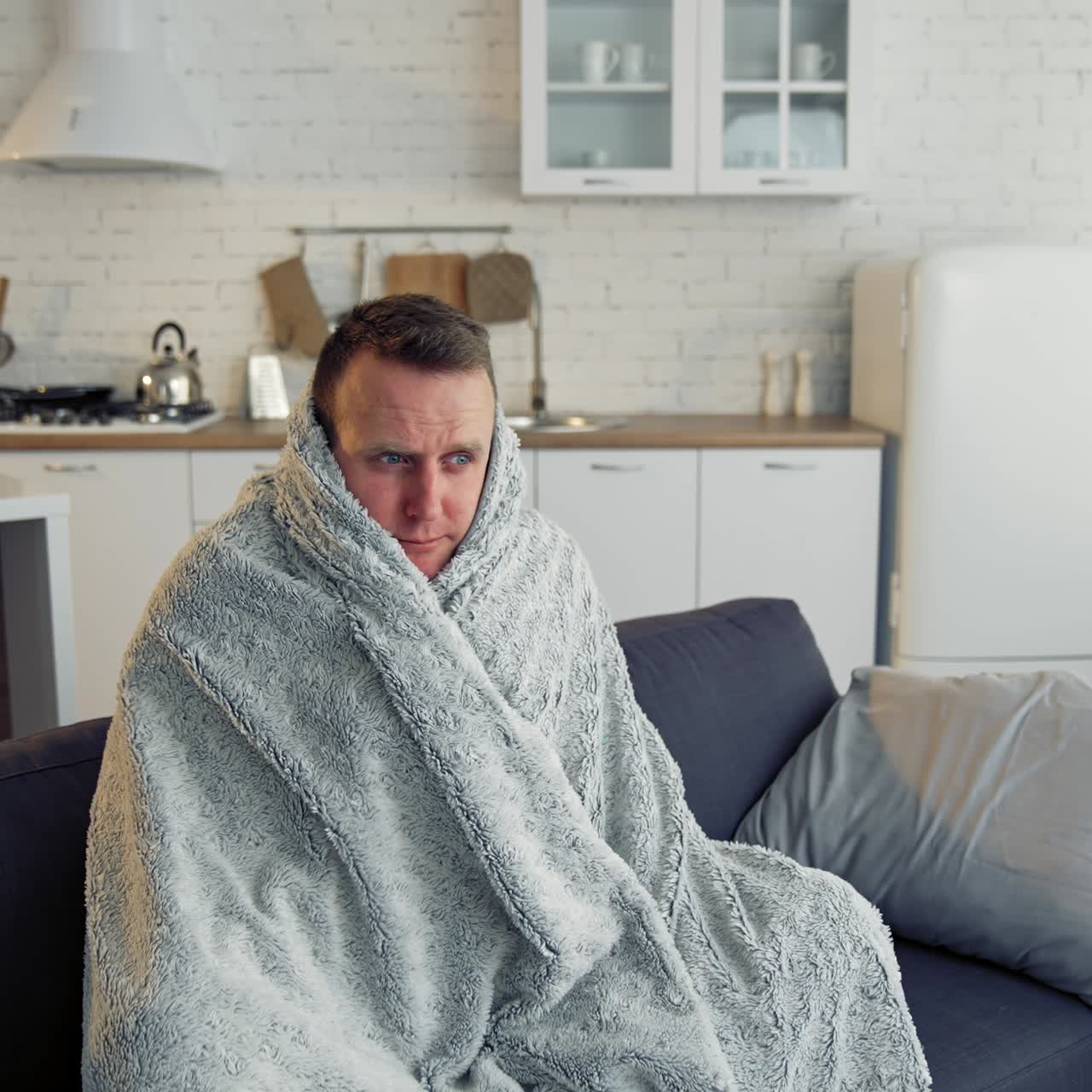 Man suffering from flu. Young sick man sitting on a sofa warming with blanket in the kitchen. Miserable man having a headache. Covid-19. Isolation at home