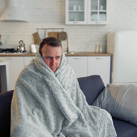 man suffering from flu. young sick man sitting on a sofa warming with blanket in the kitchen. miserable man having a headache. covid-19. isolation at home