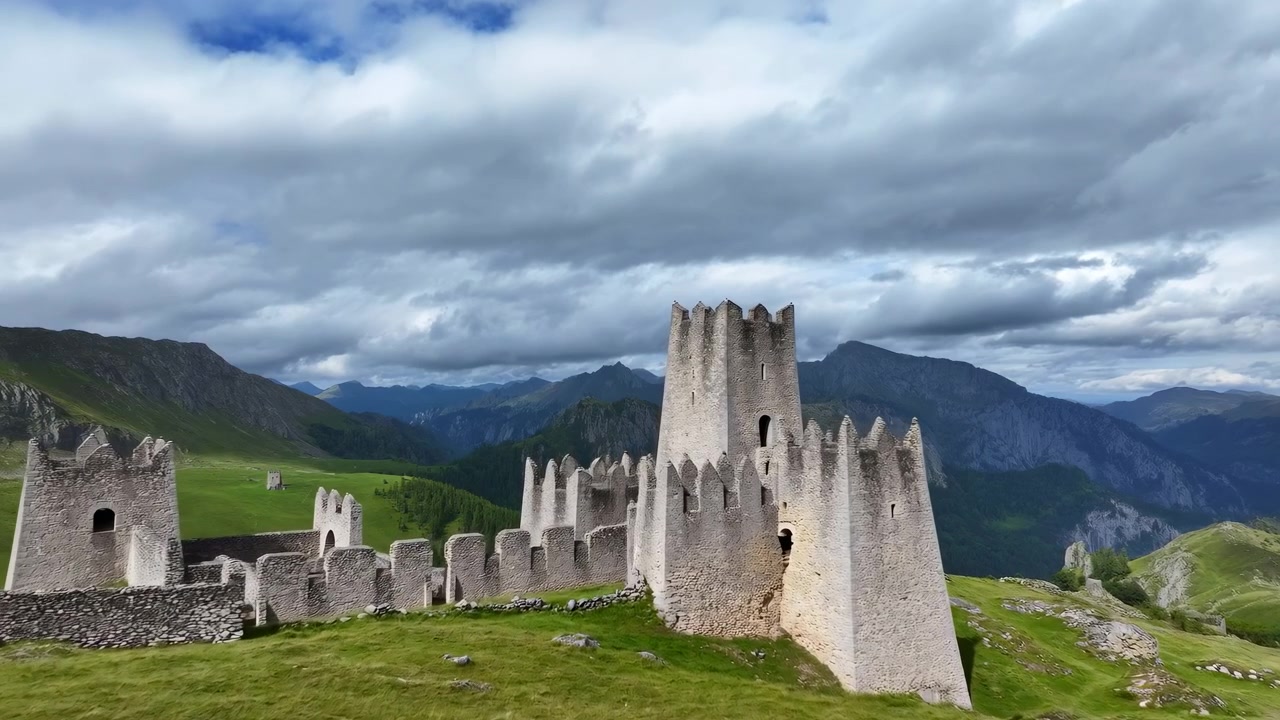 Vidsträckt naturskön utsikt över medeltida stenborgsruiner belägna på grön bergsplatå under tunga mulna moln, som visar vittrade torn, trasiga murar, steniga sluttningar och stämningsfullt historiskt landskap