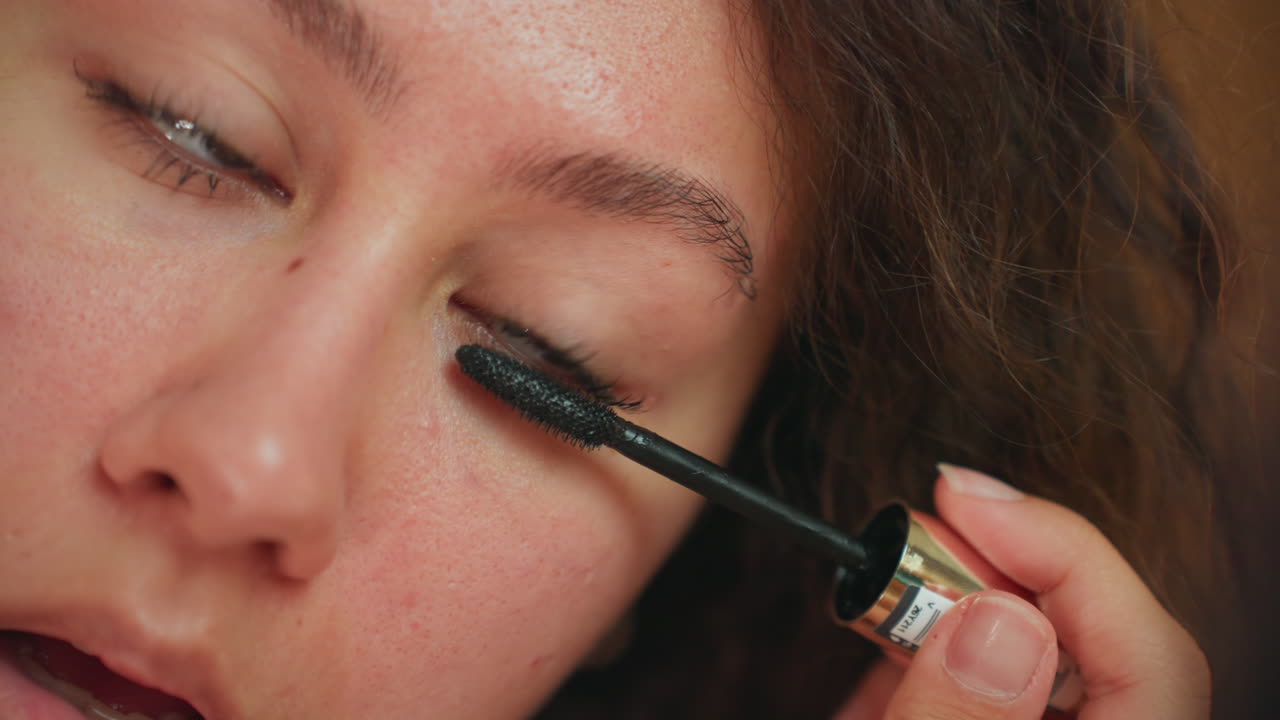 Ospite latina in una suite d'hotel, ripresa dettagliatamente mentre applica il mascara con lunghe passate su ciglia superiori e inferiori; luce luminosa dalla finestra, eleganza di una camera boutique, espressione fiduciosa e composta, pronta per un evento serale.