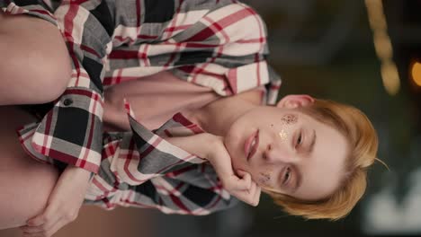 Vertical-video:-portrait-of-a-happy-mysterious-blonde-lesbian-girl-with-a-short-haircut-with-sparkles-on-her-face-in-a-checkered-pink-shirt-who-looks-at-the-camera-and-smiles