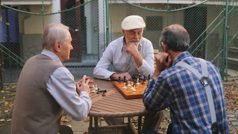 Blick-über-Den-Baum-Auf-Die-Alten-Drei-Männlichen-Freunde-Im-Ruhestand-Beim-Schachspielen-An-Der-Frischen-Luft-Im-Hof