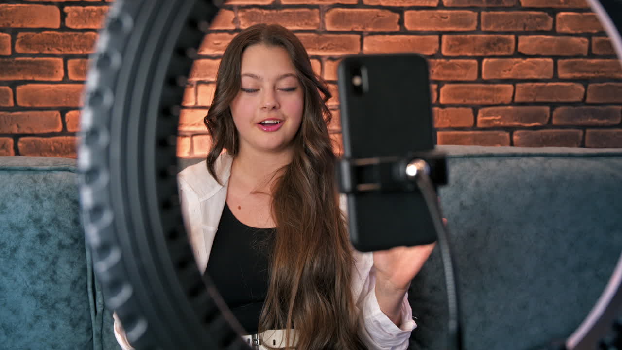 Una giovane blogger influencer sorridente che parla e si riprende con lo smartphone su un treppiede con ring light in uno studio