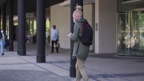 Albino-african-american-man-with-face-mask-and-dreadlocks-walking-and-using-smartphone