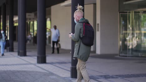 albino african american man with face mask and dreadlocks walking and using smartphone