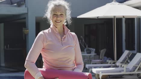 Retrato-De-Una-Feliz-Mujer-Caucásica-Mayor-Sosteniendo-Una-Estera-De-Yoga-Y-Sonriendo-En-El-Jardín,-Cámara-Lenta