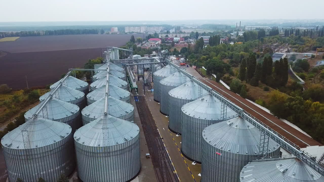 Enormous metal tanks standing in straight rows. Railways dividing the plant territory. Cityscape and farmlands at backdrop.