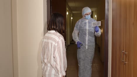 man in a hazmat suit and a face mask standing in a hallway, holding up a sign that says 'don't miss your shot to stop covid-19' and talking to a woman who is wearing a mask and clapping