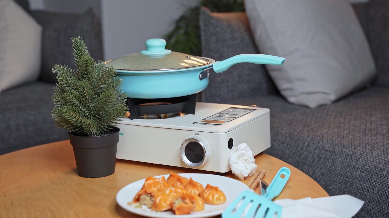 A light blue pan sits on a white portable gas stove cooking orange dumplings. Plated dim sum, a spatula, and a small pine plant rest on a wooden table in a modern indoor living room setting.