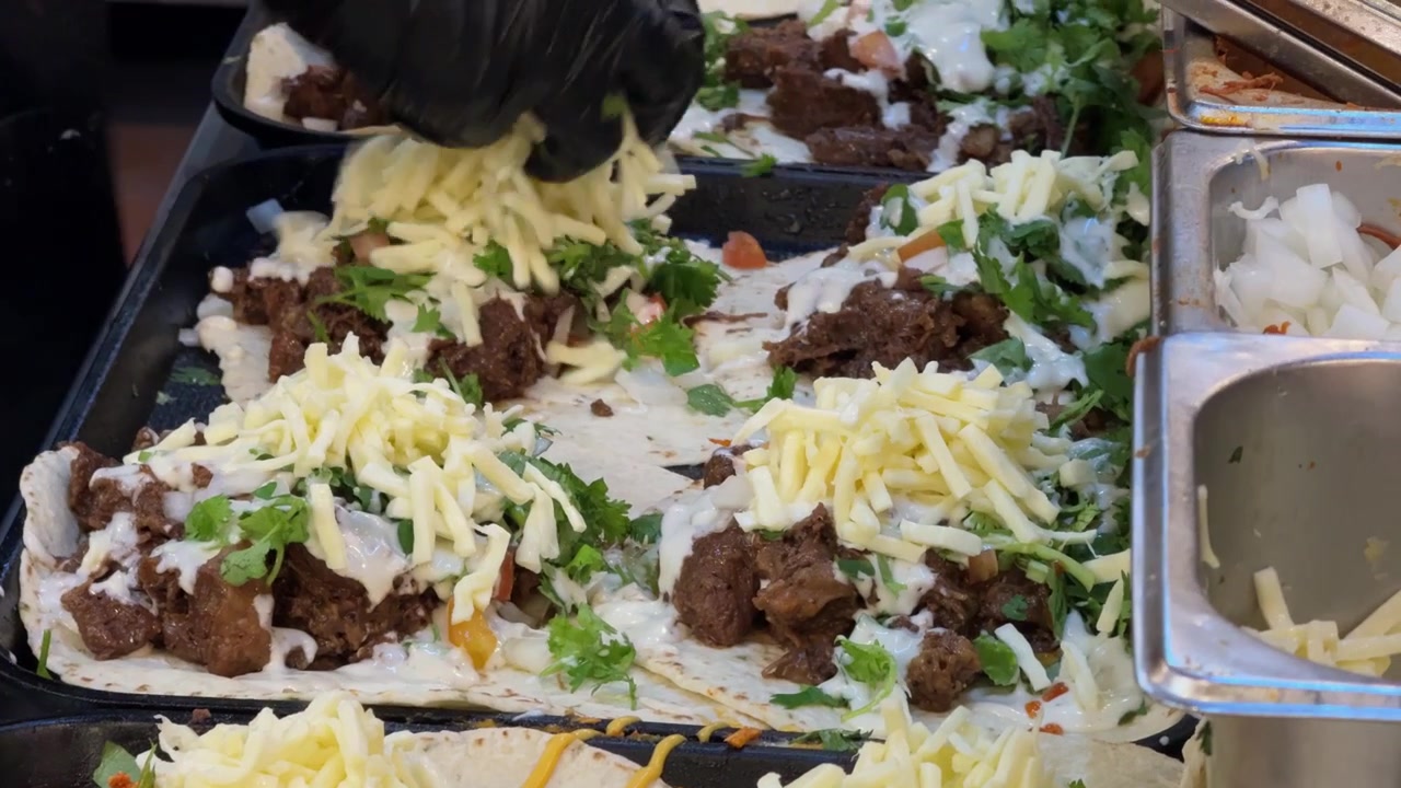 Filmato ravvicinato di un venditore ambulante che prepara numerosi tacos e wrap freschi su una piastra calda. Le tortillas sono riempite con carne, formaggio, verdure, erbe aromatiche e salse durante la frenetica preparazione di cibo da strada.
