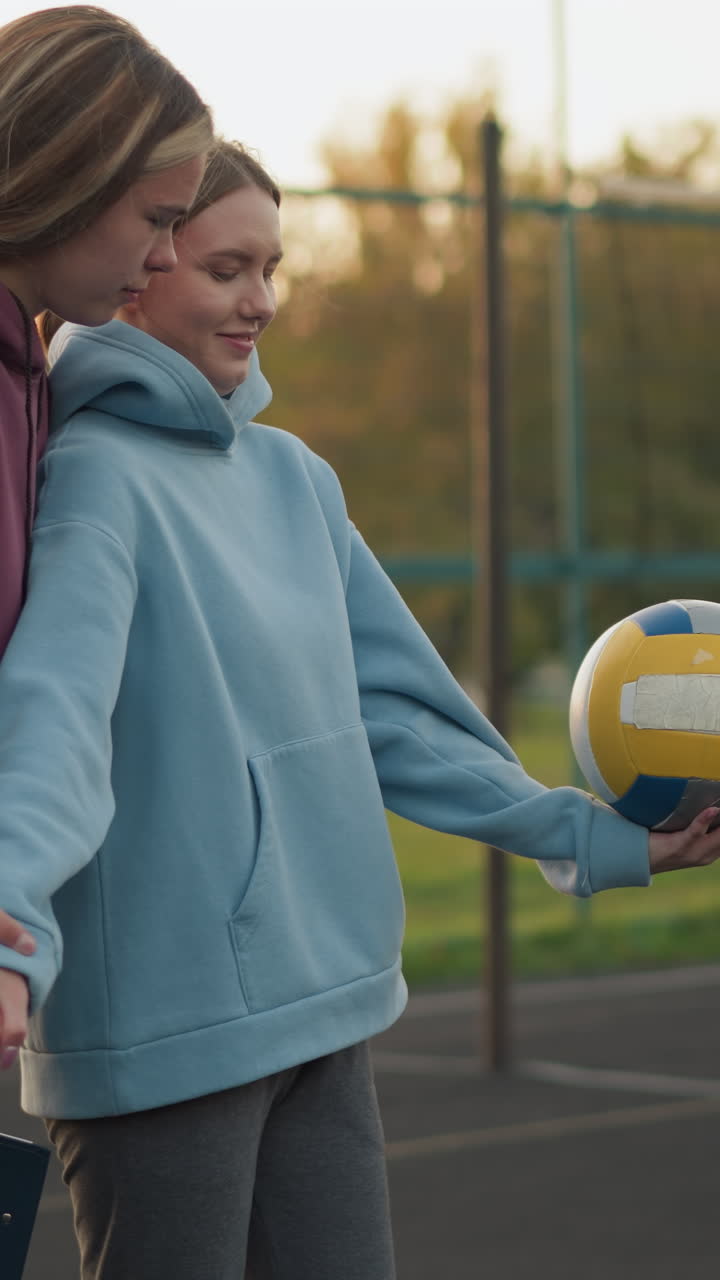vista lateral de un instructor de voleibol enseñando a un principiante cómo servir, ambos sosteniendo la pelota con enfoque y determinación, de pie en la cancha al aire libre con el edificio y la red en el fondo