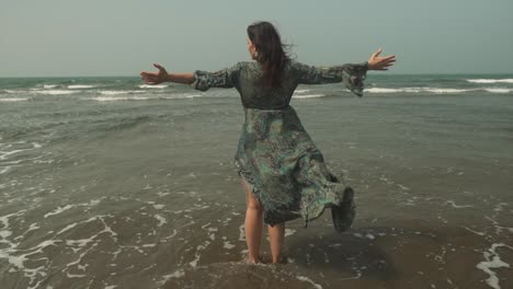 caucasian woman stands with open arms, dress blows in ocean breeze, slowmo