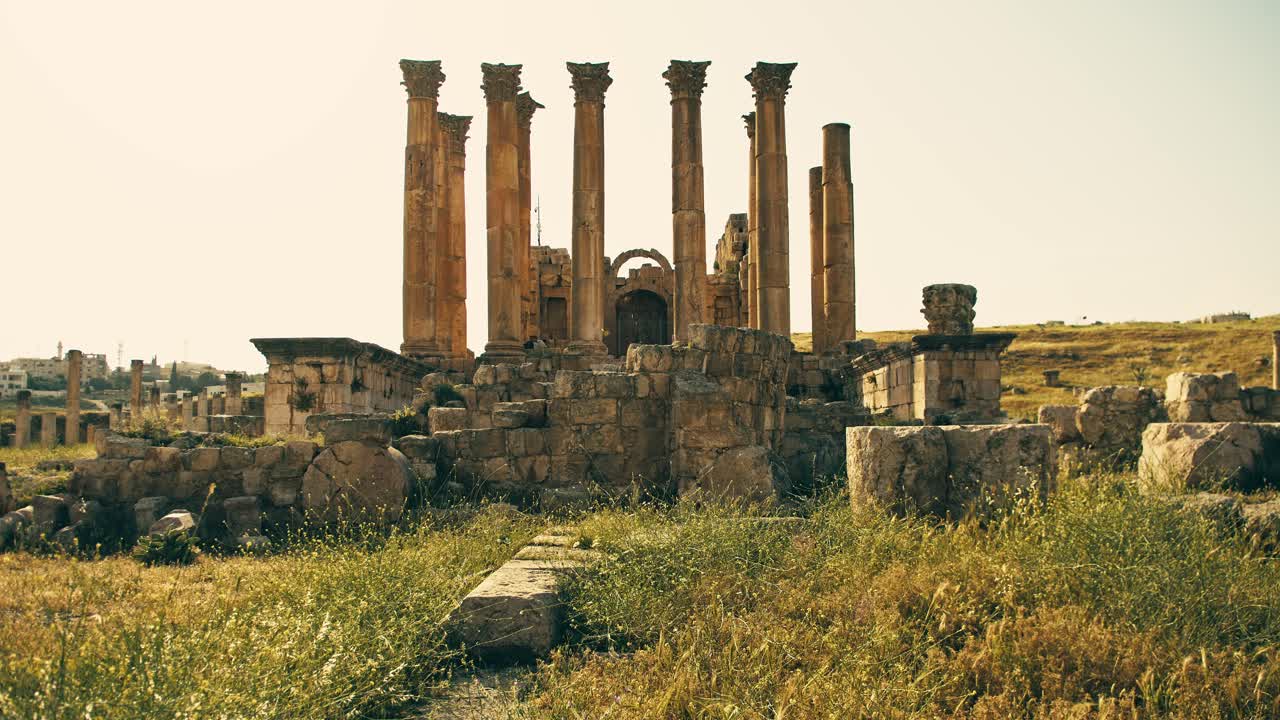 Campo lungo di antiche rovine romane a Jerash, in Giordania, con alte colonne di pietra ed erba selvatica che si muove dolcemente in primo piano. Luce calda del giorno, composizione cinematografica, atmosfera storica senza tempo
