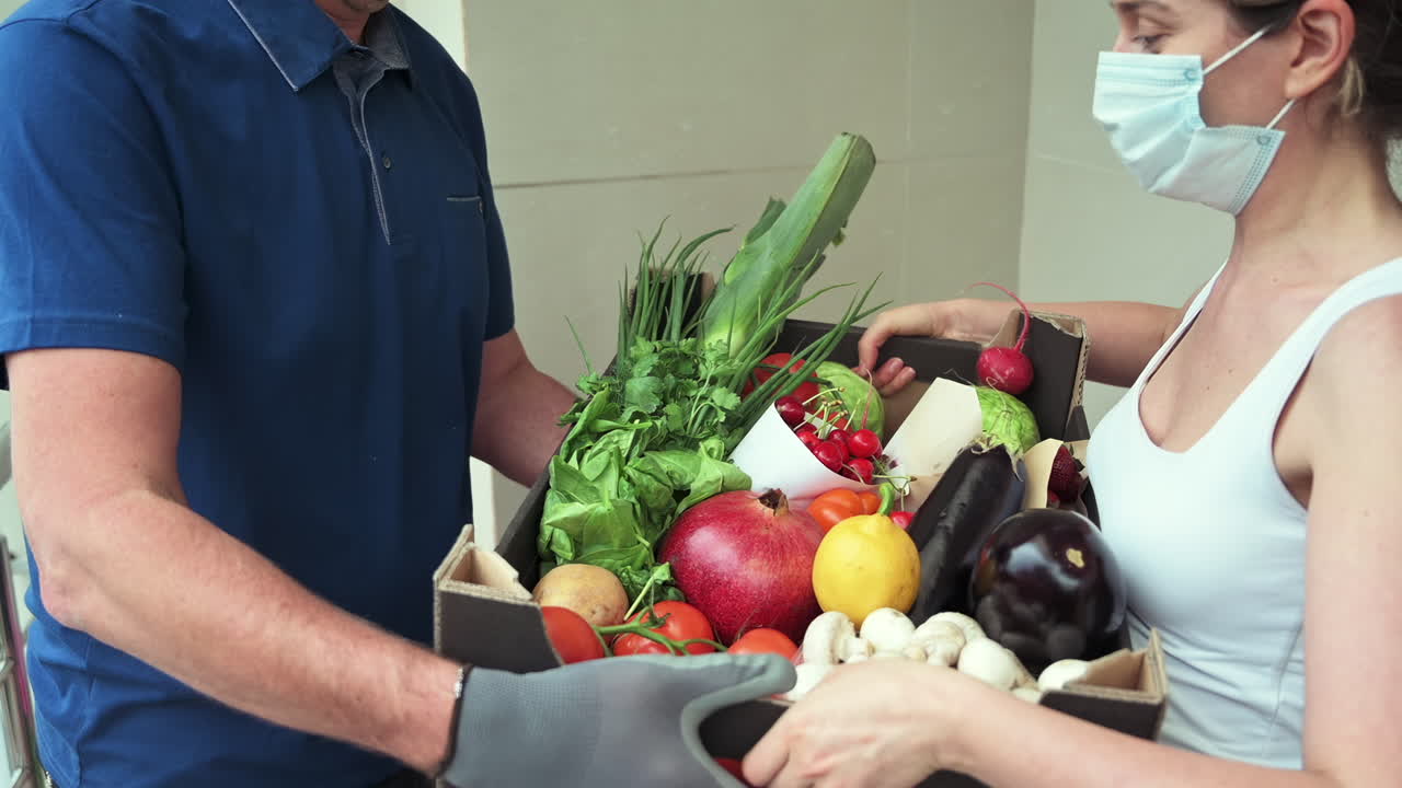 Woman receiving a box of fresh fruits and vegetables from a masked delivery man