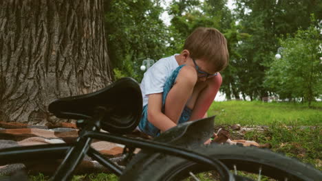 a young child squats under a large tree, deep in thought, a bicycle lies on the grass beside him, with trees in the background