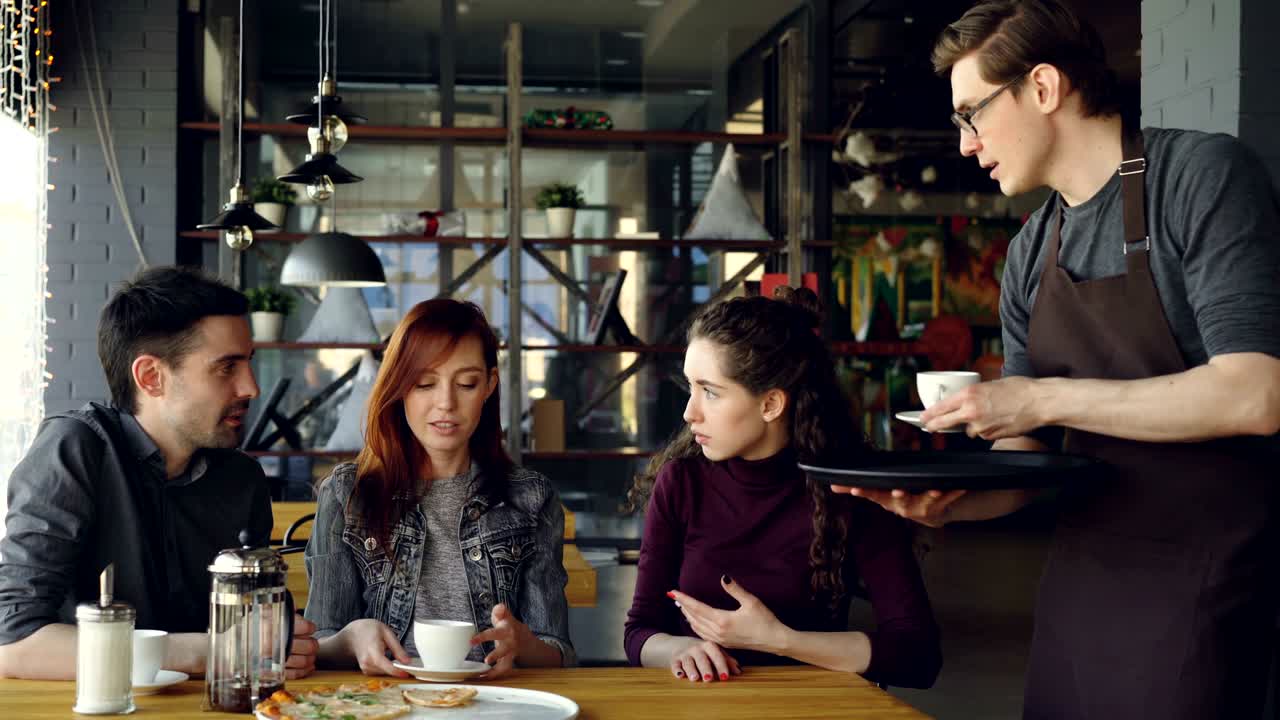 옷을 입은 친절한 웨이터는 테이블에 앉은 카페에서 사교하는 젊은이들에게 음료 컵을 제공합니다. 음식과 음료 산업, 외식, 친구와 우정 개념을 만나는 것.