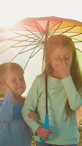 boy and girl protected by rainbow parasol. siblings unhappy because of drops on face standing beneath umbrella shielding from playful raindrops and sun