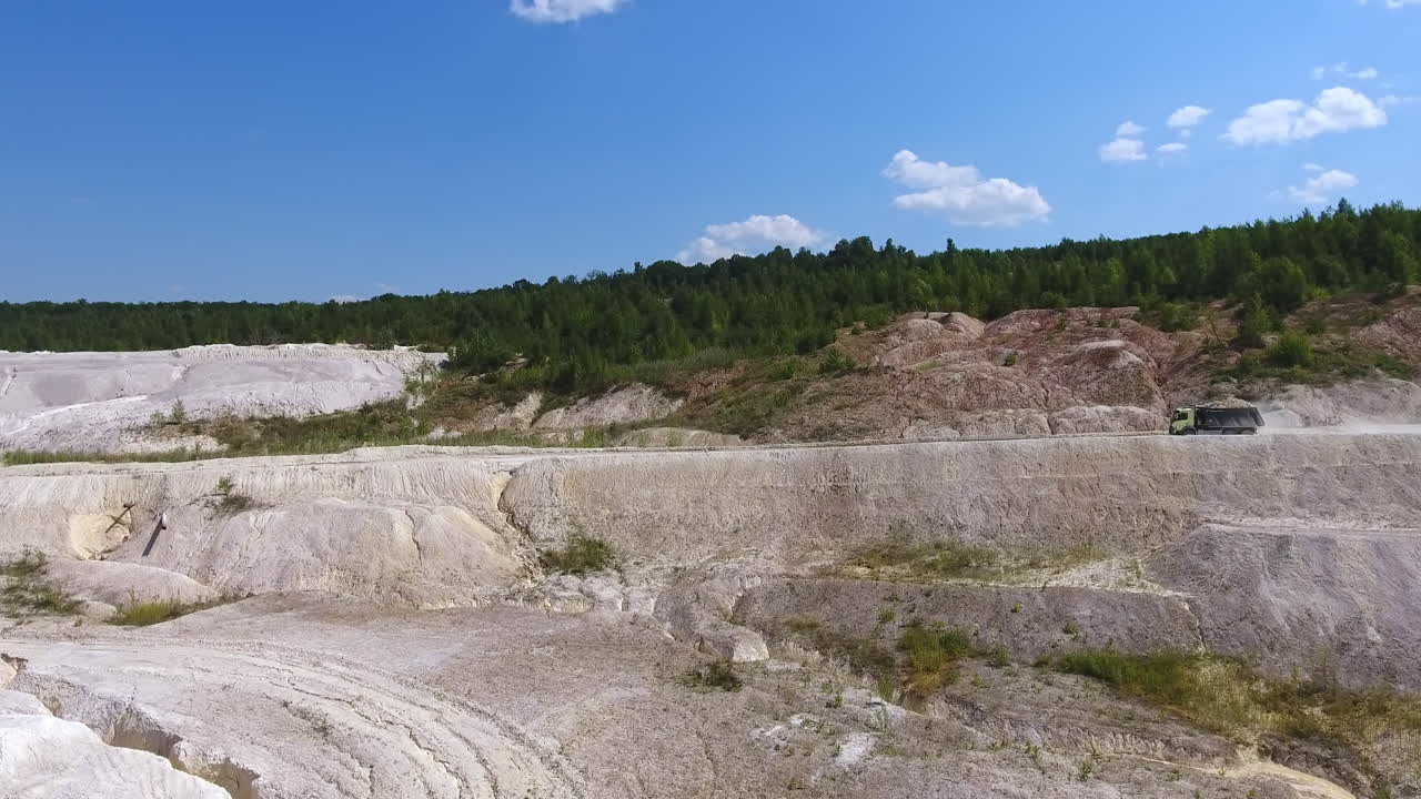 Truck loaded full with kaolinite heading to the modern plant. Uneven ground of white clay contrasting with green forest and blue sky.