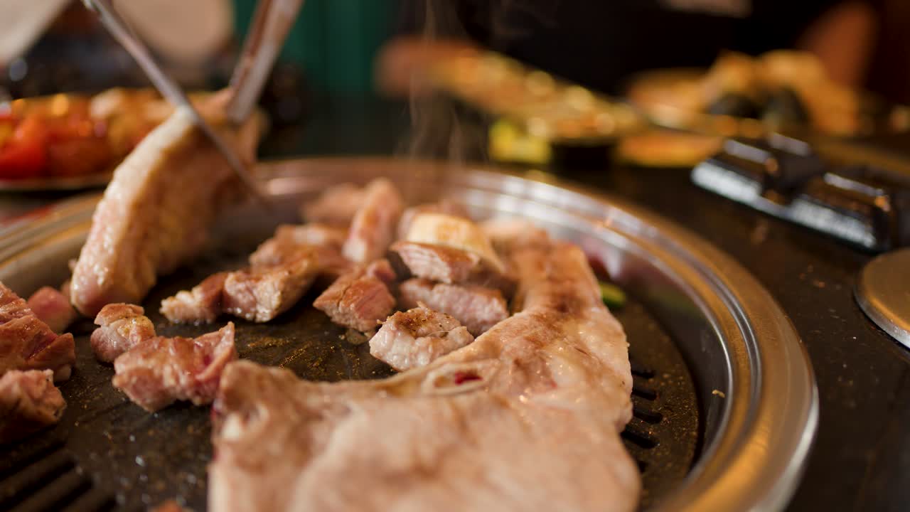 Close-up of pork belly grilling, tongs turning meat, indoor restaurant, warm lighting, shallow focus