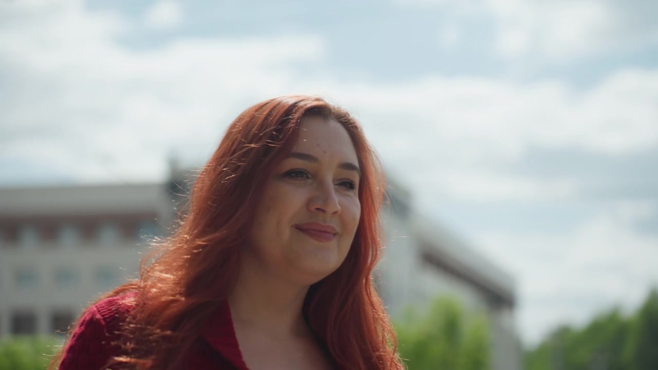 Nærbillede af kvinde med langt rødt hår iført en bordeaux striktrøje, der holder og kigger på en smartphone udendørs. Hendes udtryk er roligt, mens kvinden interagerer med enheden mod en urban baggrund.