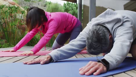 Happy-diverse-couple-doing-yoga,-stretching-on-yoga-mat-at-terrace