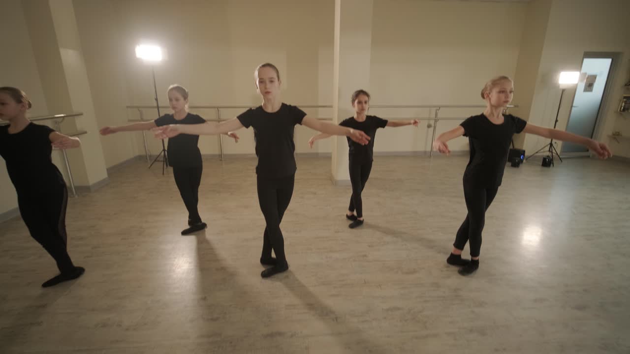 un grupo de jóvenes estudiantes de ballet con ropa de baile negra practicando posiciones en un amplio estudio de ballet con pisos de madera y barras montadas en la pared. expresiones enfocadas y movimientos sincronizados.