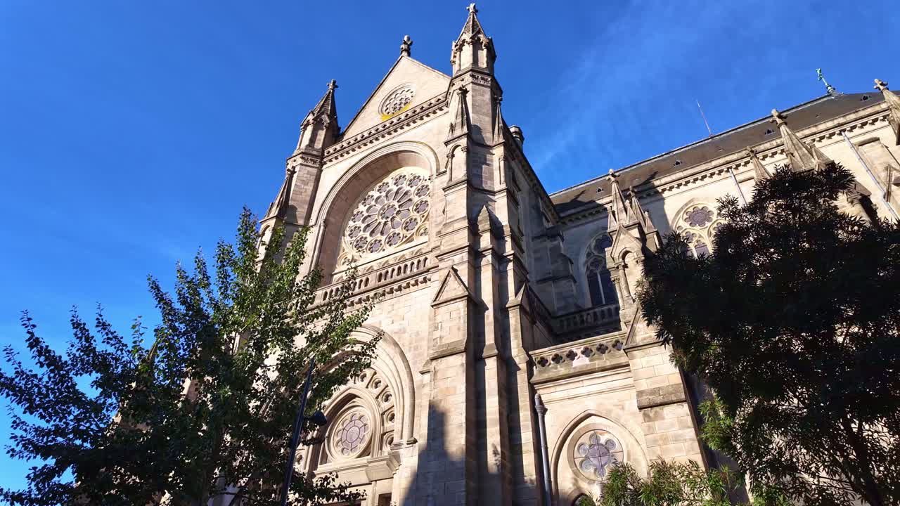 Low angle ground shot of the Basilica of Saint-Aubin Notre-Dame de Bonne-Nouvelle in Rennes, Saint-Anne square, showing the rose window, facade, and surrounding trees under bright sunlight