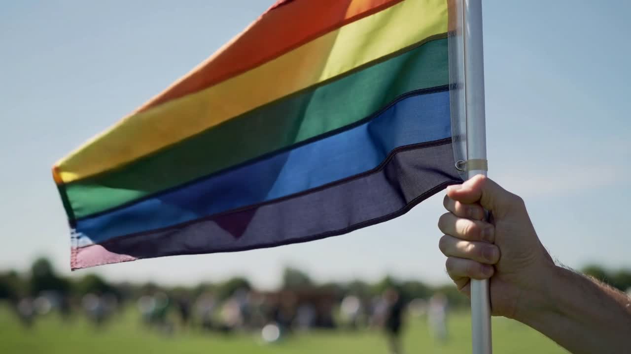 Holder's right hand gripping pole, breeze causing rainbow flag rippling, displaying pride at rally