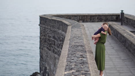 on a classic european square near the ocean, a young mother walks with her baby son, observing the waves and smiling warmly at each other