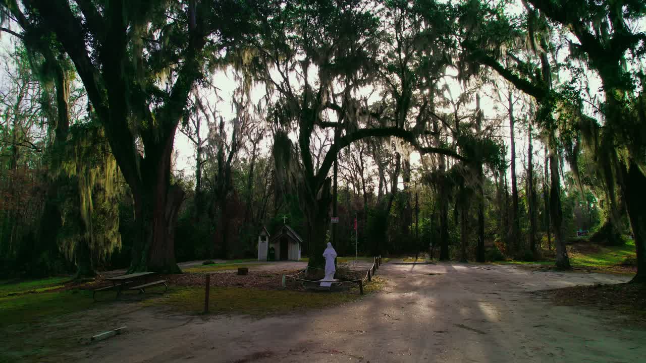 Historic tiny chapel surrounded by majestic oak trees and Spanish moss, featuring a serene dirt path and memorial statue in a tranquil forest setting.