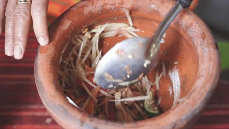 cooking som tam series: slo mo street food vendor's hand mixing som tam or thai spicy green papaya salad ingredients in mortar using ladle before serving at street food stall