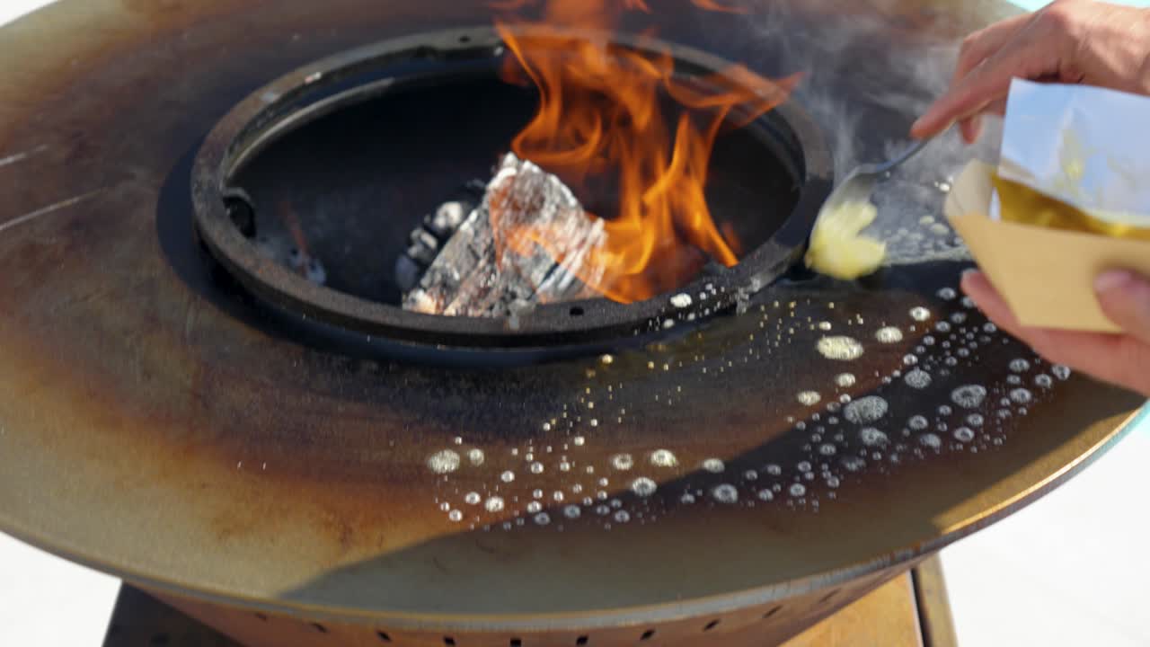 close-up of a barbecue with the flame and embers in the centre and the cooking plate around it, a man's hand melting butter on the plate