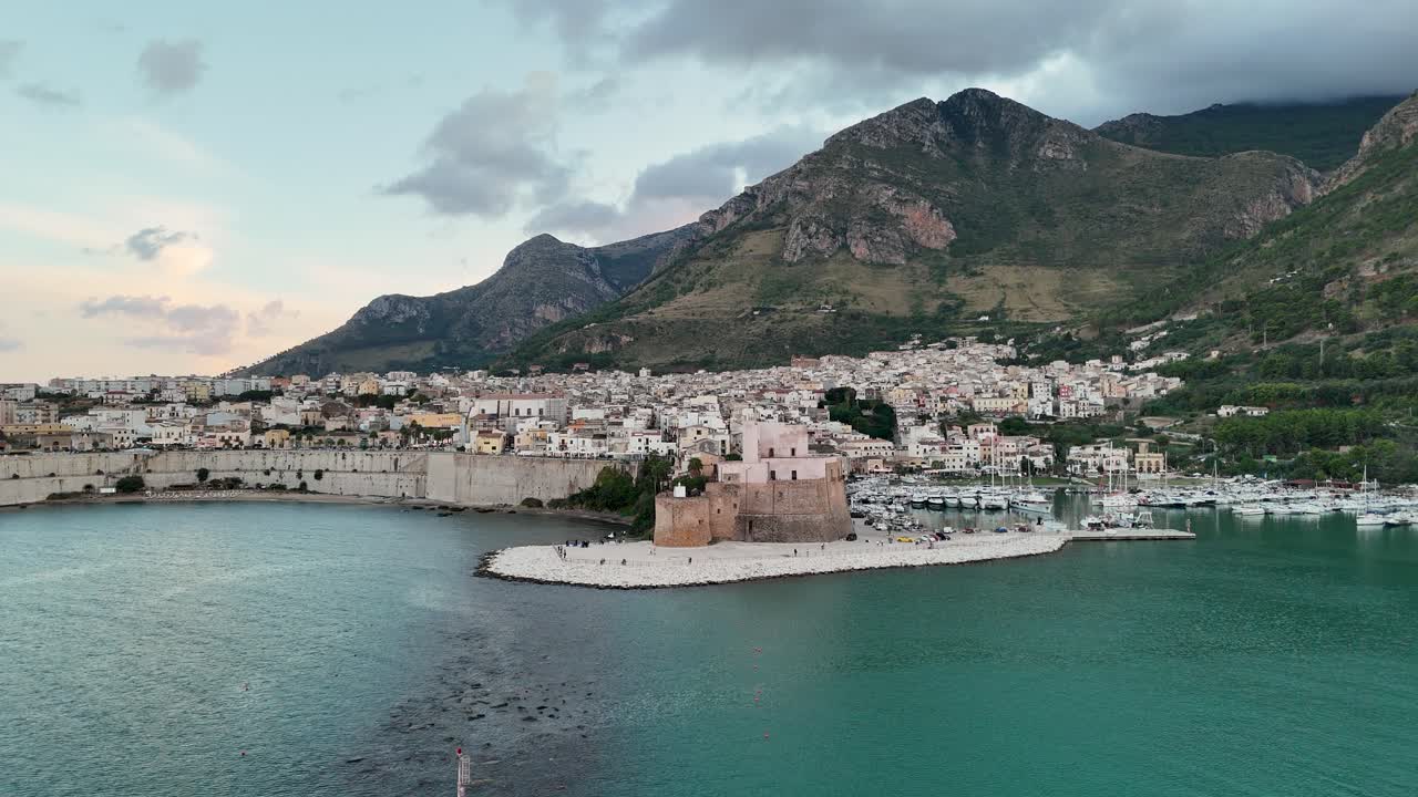 Aerial drone fly-in toward the harbor and historic castle of Castellammare del Golfo, Sicily. Scenic coastal town framed by turquoise sea and dramatic mountains