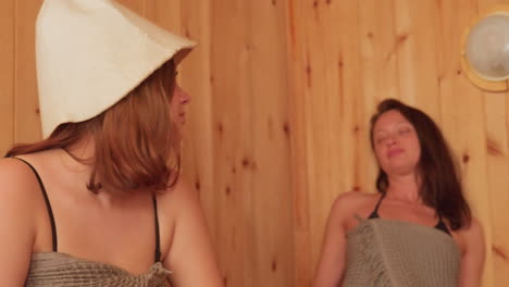 Two women in towels smiling at each other in a cozy sauna, sharing a moment of happiness and relaxation, they are seated on a wooden bench with a serene wooden backdrop, enjoying their time