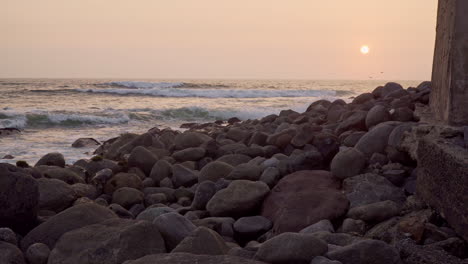 Static-shot-of-the-sun-setting-over-the-horizon-with-a-rocky-beach-in-the-foreground-along-the-coast-in-San-Bartolo,-Lima,-Peru